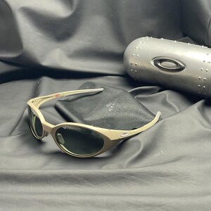 Puede incluir: Gafas de sol Oakley color beige con lentes oscuras, un estuche negro y un estuche de metal plateado. Las gafas de sol tienen un dise&ntilde;o de montura curvada. El estuche negro tiene el logotipo de Oakley. El estuche plateado tiene un dise&ntilde;o remachado.