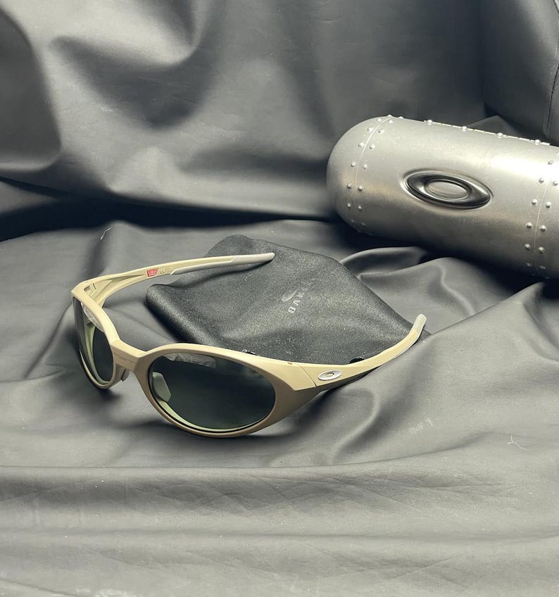 Puede incluir: Gafas de sol Oakley color beige con lentes oscuras, un estuche negro y un estuche de metal plateado. Las gafas de sol tienen un dise&ntilde;o de montura curvada. El estuche negro tiene el logotipo de Oakley. El estuche plateado tiene un dise&ntilde;o remachado.