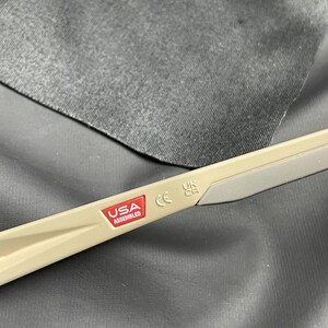 Puede incluir: Primer plano de la patilla de unas gafas. La patilla es de color beige con una punta gris. Una pegatina roja y blanca dice "USA ASSEMBLED". Tambi&eacute;n son visibles las letras "CE" y "UK CA".
