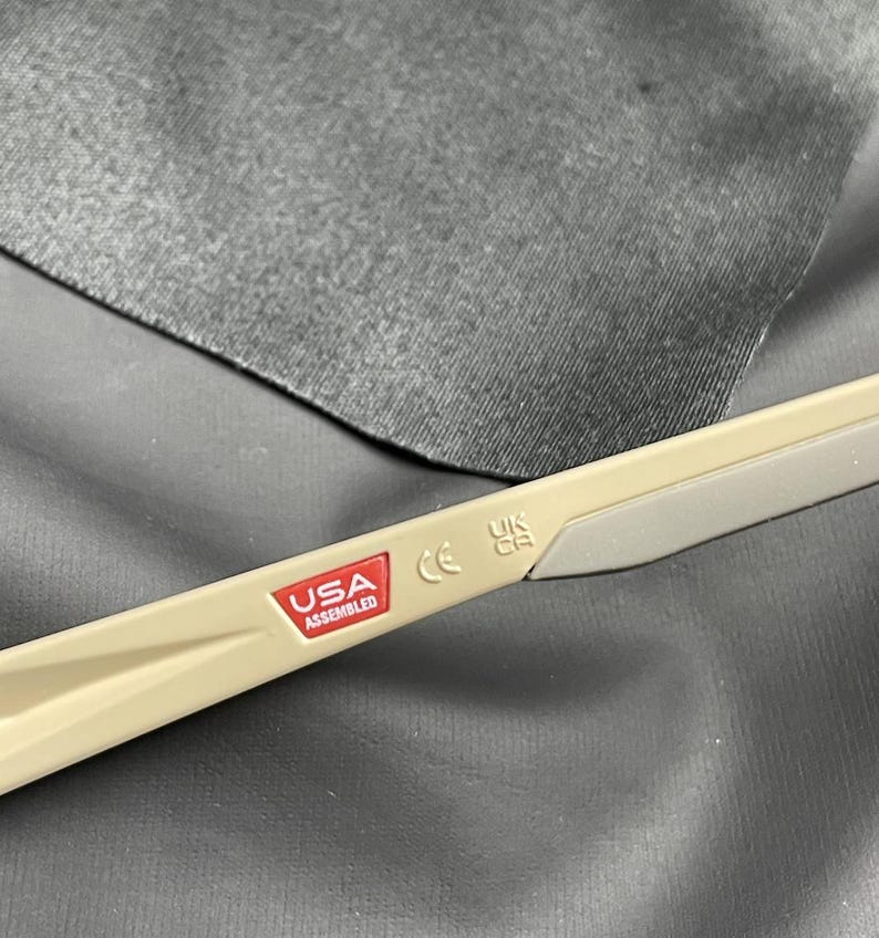 Puede incluir: Primer plano de la patilla de unas gafas. La patilla es de color beige con una punta gris. Una pegatina roja y blanca dice "USA ASSEMBLED". Tambi&eacute;n son visibles las letras "CE" y "UK CA".