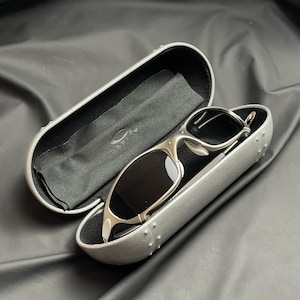May include: Silver-framed sunglasses with dark lenses rest in an open, gray case. The case is lined with black fabric and includes a cleaning cloth. The Oakley logo is visible on the case and cloth. The sunglasses have a sleek, modern design.