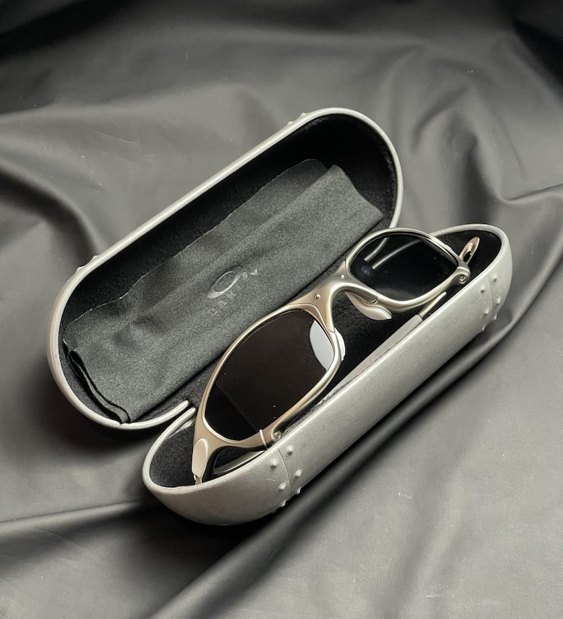 May include: Silver-framed sunglasses with dark lenses rest in an open, gray case. The case is lined with black fabric and includes a cleaning cloth. The Oakley logo is visible on the case and cloth. The sunglasses have a sleek, modern design.