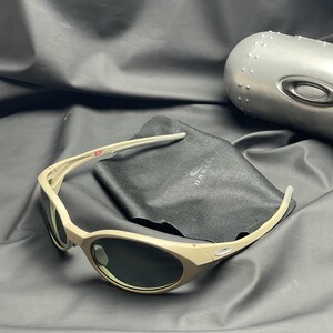 Puede incluir: Gafas de sol color beige con lentes oscuras, una funda de microfibra negra y un estuche plateado. Las gafas tienen un dise&ntilde;o curvo y el nombre de la marca es visible en la funda. El estuche es met&aacute;lico y tiene una superficie texturizada.