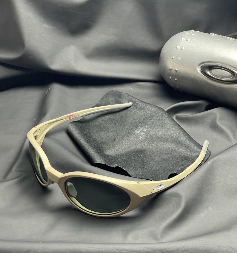 Puede incluir: Gafas de sol color beige con lentes oscuras, una funda de microfibra negra y un estuche plateado. Las gafas tienen un dise&ntilde;o curvo y el nombre de la marca es visible en la funda. El estuche es met&aacute;lico y tiene una superficie texturizada.