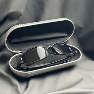 May include: Black sunglasses with dark lenses rest inside an open, silver-colored case with a black interior. The sunglasses have a sleek, modern design. The case is slightly open, revealing the sunglasses.