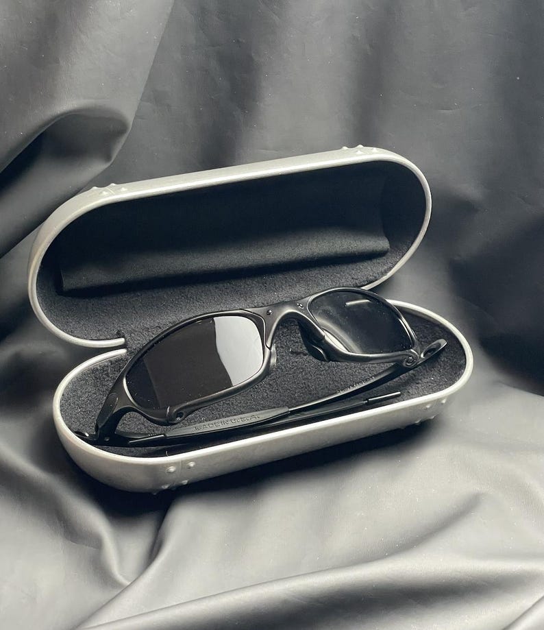 May include: Black sunglasses with dark lenses rest inside an open, silver-colored case with a black interior. The sunglasses have a sleek, modern design. The case is slightly open, revealing the sunglasses.