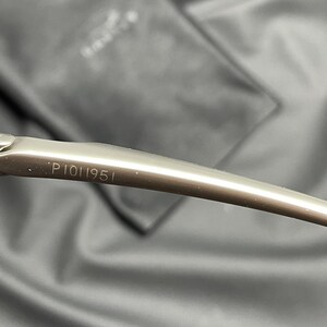 May include: Close-up of a silver-toned eyeglass arm with the number "P1011951" etched on the surface. The arm curves gently, and the background is a blurred, dark gray fabric.