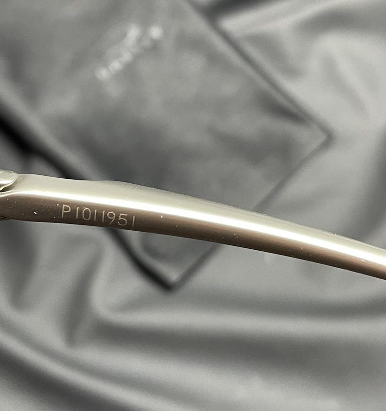 May include: Close-up of a silver-toned eyeglass arm with the number "P1011951" etched on the surface. The arm curves gently, and the background is a blurred, dark gray fabric.