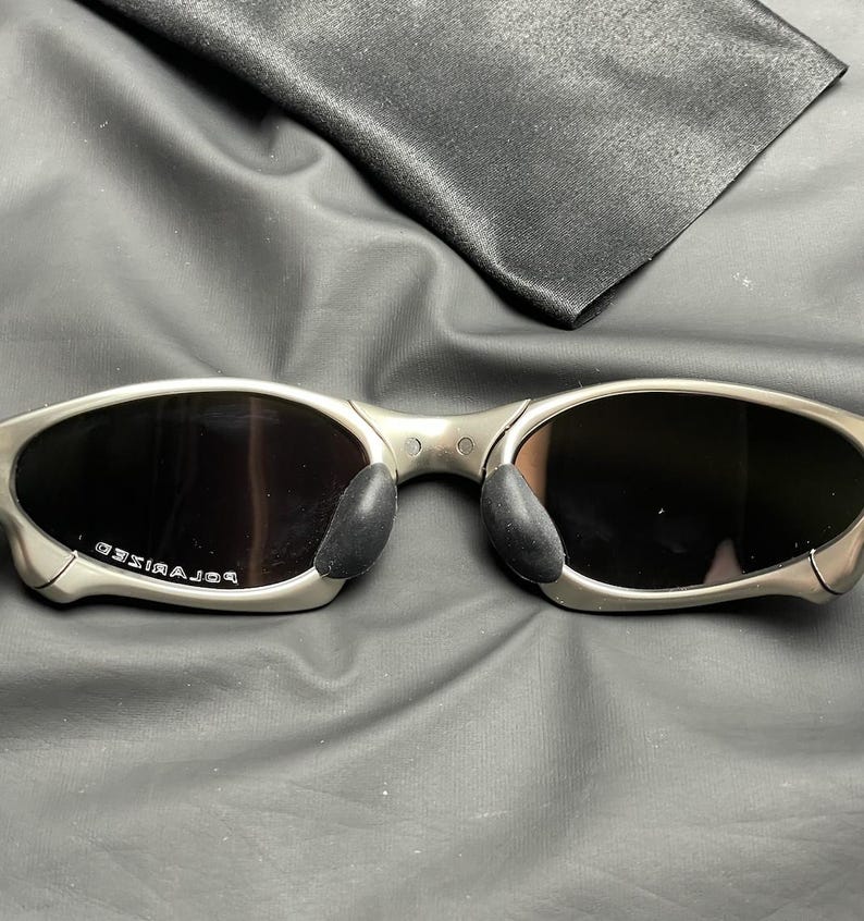 May include: Silver-framed sunglasses with dark lenses and black nose pads. The word "POLARIZED" is printed on the left lens. A black microfiber cleaning cloth is in the background.