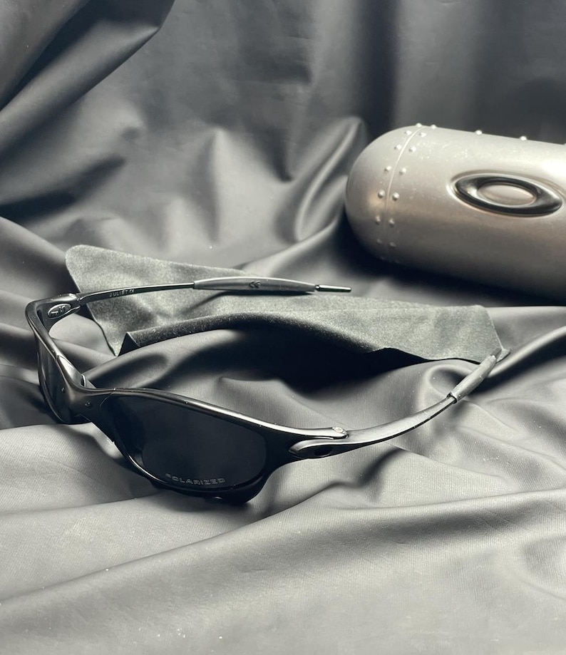 May include: Black polarized sunglasses with a sleek, modern design. The sunglasses have a curved frame and dark lenses. A gray case and a black microfiber cloth are also present.