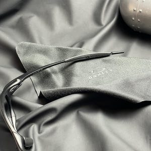 May include: Black Oakley sunglasses with a sleek, curved design. The sunglasses are resting on a dark gray microfiber cloth with the Oakley logo. The frame has the text "JULIET CE".