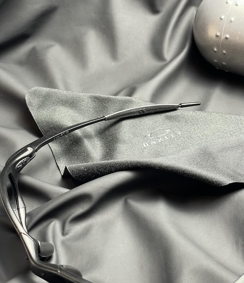 May include: Black Oakley sunglasses with a sleek, curved design. The sunglasses are resting on a dark gray microfiber cloth with the Oakley logo. The frame has the text "JULIET CE".