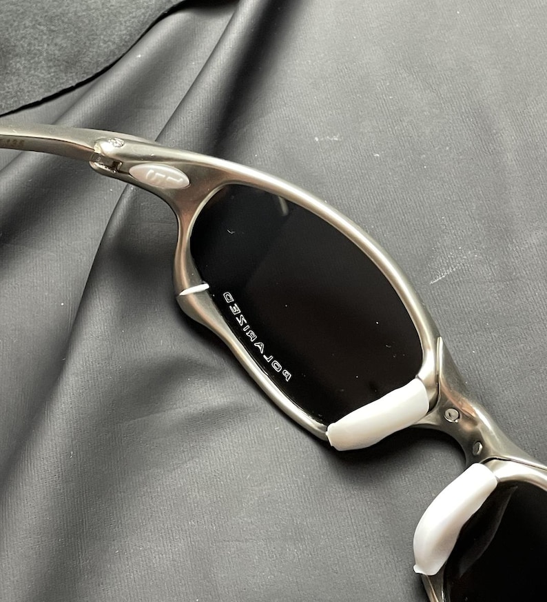 May include: Silver-framed sunglasses with black polarized lenses. The sunglasses have white nose pads and the word "POLARIZED" is printed on the lens. The frame is a metallic silver color.