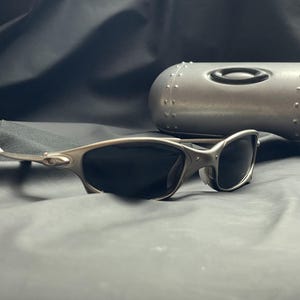 May include: Silver-framed sunglasses with dark lenses rest on a dark gray surface. A matching silver case with the Oakley logo is in the background. The sunglasses have a sleek, modern design.