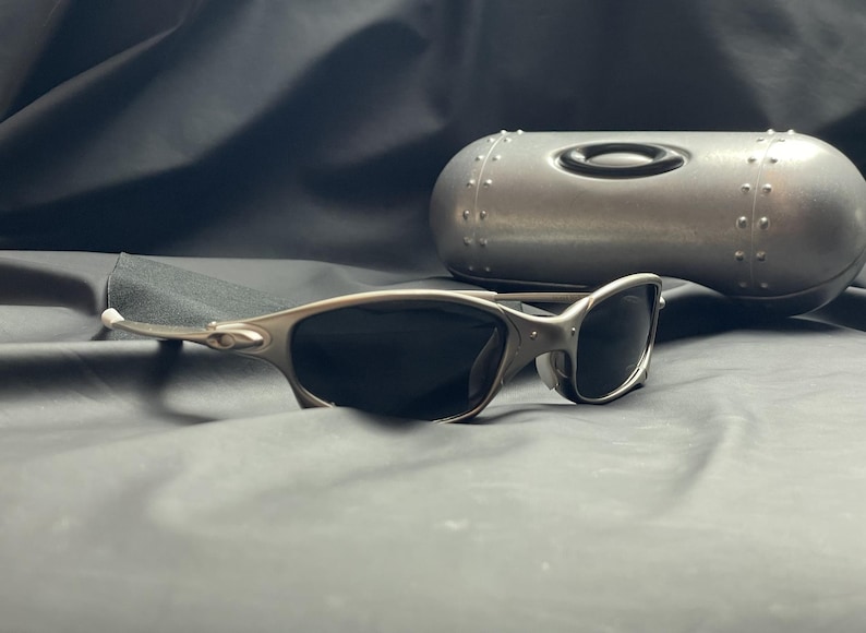 May include: Silver-framed sunglasses with dark lenses rest on a dark gray surface. A matching silver case with the Oakley logo is in the background. The sunglasses have a sleek, modern design.