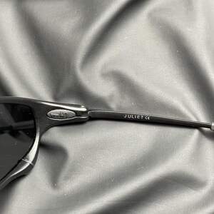May include: Close-up of black sunglasses with the text "JULIET CE" printed on the temple. The sunglasses have a sleek, modern design with dark lenses and a matte finish. The frame appears to be made of a durable material.