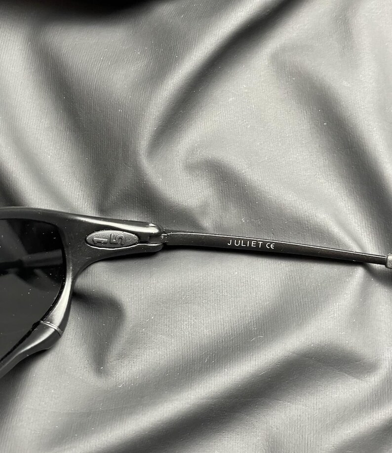 May include: Close-up of black sunglasses with the text "JULIET CE" printed on the temple. The sunglasses have a sleek, modern design with dark lenses and a matte finish. The frame appears to be made of a durable material.