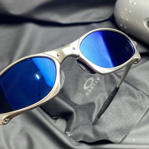 May include: Silver-framed sunglasses with blue lenses. The sunglasses have a sleek, modern design with a metal frame and a black temple tip. The Oakley logo is visible on the lens cloth.