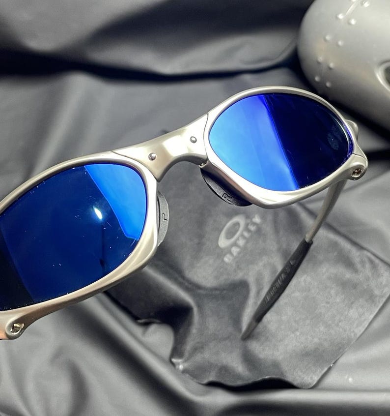 May include: Silver-framed sunglasses with blue lenses. The sunglasses have a sleek, modern design with a metal frame and a black temple tip. The Oakley logo is visible on the lens cloth.