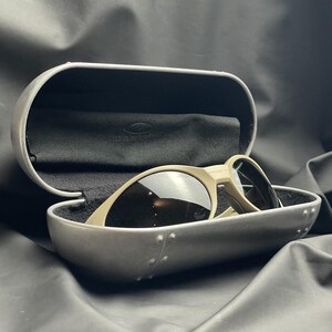 Puede incluir: Unas gafas de sol con montura beige claro y lentes oscuras se encuentran dentro de un estuche plateado abierto con interior negro. El estuche presenta un dise&ntilde;o remachado. Tambi&eacute;n se ve un pa&ntilde;o negro.
