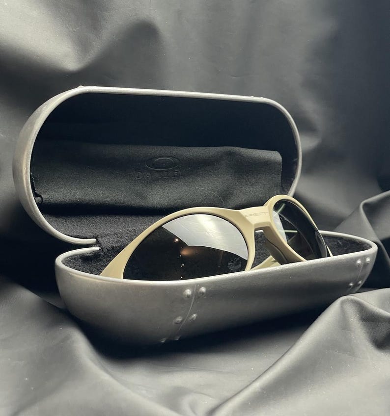 Puede incluir: Unas gafas de sol con montura beige claro y lentes oscuras se encuentran dentro de un estuche plateado abierto con interior negro. El estuche presenta un dise&ntilde;o remachado. Tambi&eacute;n se ve un pa&ntilde;o negro.
