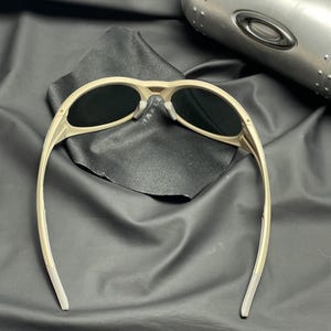 Puede incluir: Gafas de sol beige con lentes oscuras, sobre una tela negra. Las gafas de sol tienen una montura curvada y un dise&ntilde;o moderno. Una funda met&aacute;lica es visible en el fondo.