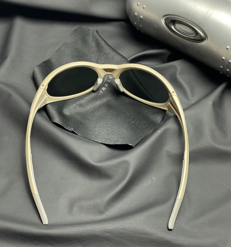 Puede incluir: Gafas de sol beige con lentes oscuras, sobre una tela negra. Las gafas de sol tienen una montura curvada y un dise&ntilde;o moderno. Una funda met&aacute;lica es visible en el fondo.
