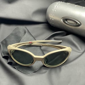 Puede incluir: Gafas de sol color beige con lentes oscuras sobre una tela negra. Las gafas tienen una montura curvada y un dise&ntilde;o elegante. Un estuche plateado en el fondo. Un accesorio de moda.
