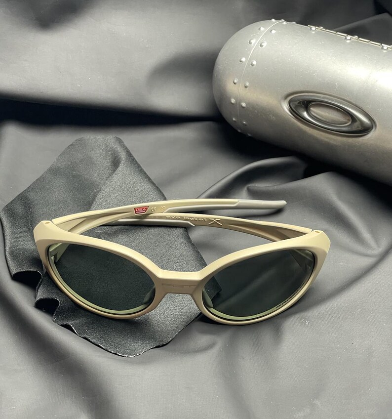 Puede incluir: Gafas de sol color beige con lentes oscuras sobre una tela negra. Las gafas tienen una montura curvada y un dise&ntilde;o elegante. Un estuche plateado en el fondo. Un accesorio de moda.