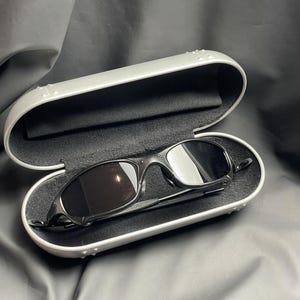 May include: Black sunglasses with dark lenses rest inside an open, silver-colored case. The case has a black, soft interior. The sunglasses have a sleek, modern design. The case is on a dark, textured surface.