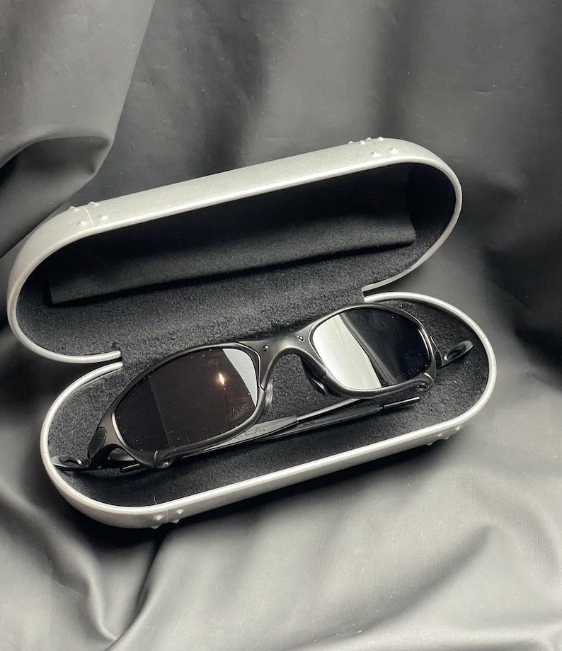May include: Black sunglasses with dark lenses rest inside an open, silver-colored case. The case has a black, soft interior. The sunglasses have a sleek, modern design. The case is on a dark, textured surface.