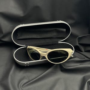 Puede incluir: Gafas de sol ojo de gato blanquecinas con lentes oscuras dentro de un estuche plateado abierto con interior negro. El estuche est&aacute; sobre una superficie negra. Las gafas tienen un dise&ntilde;o retro.