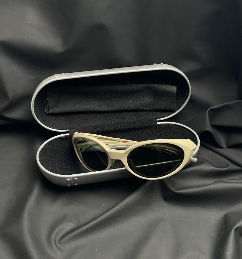 Puede incluir: Gafas de sol ojo de gato blanquecinas con lentes oscuras dentro de un estuche plateado abierto con interior negro. El estuche est&aacute; sobre una superficie negra. Las gafas tienen un dise&ntilde;o retro.