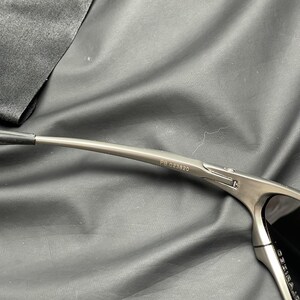 May include: Close-up of a pair of sunglasses with a metallic gray frame. The text "PB 023820" is printed on the temple arm. The lenses are dark, and the sunglasses are set against a dark background.