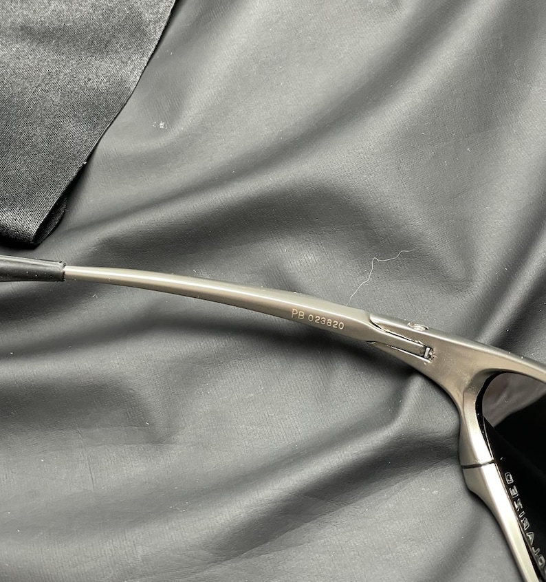 May include: Close-up of a pair of sunglasses with a metallic gray frame. The text "PB 023820" is printed on the temple arm. The lenses are dark, and the sunglasses are set against a dark background.