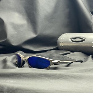 May include: Silver-framed sunglasses with blue lenses rest on a black cloth. The sunglasses have a sleek, modern design. A silver case with the brand logo is in the background.