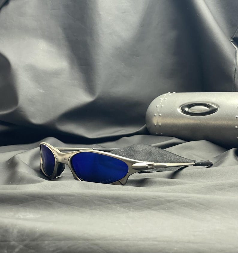 May include: Silver-framed sunglasses with blue lenses rest on a black cloth. The sunglasses have a sleek, modern design. A silver case with the brand logo is in the background.