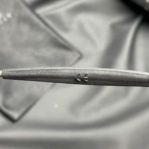 May include: Close-up of a dark gray eyeglass arm with a CE marking. The arm is textured and appears to be made of a matte material. The background is a blurred, dark fabric.
