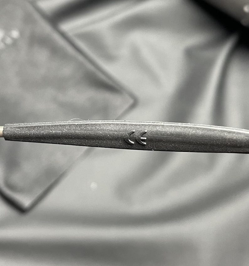 May include: Close-up of a dark gray eyeglass arm with a CE marking. The arm is textured and appears to be made of a matte material. The background is a blurred, dark fabric.