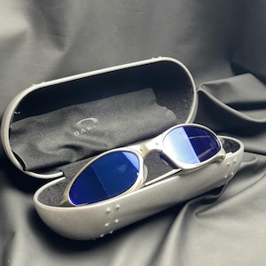 May include: Silver-framed sunglasses with blue lenses rest in a matching case. The case is open, revealing a black interior and a black cleaning cloth. The sunglasses have a sleek, modern design, perfect for outdoor activities.