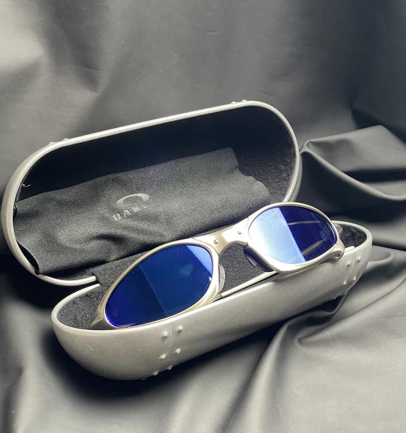 May include: Silver-framed sunglasses with blue lenses rest in a matching case. The case is open, revealing a black interior and a black cleaning cloth. The sunglasses have a sleek, modern design, perfect for outdoor activities.