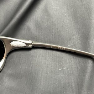 May include: Close-up of a pair of silver sunglasses with black lenses. The temple arm is visible, with the word "JULIET CE" printed on it. The sunglasses have a modern, sporty design.