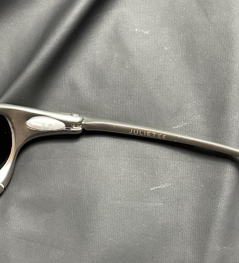 May include: Close-up of a pair of silver sunglasses with black lenses. The temple arm is visible, with the word "JULIET CE" printed on it. The sunglasses have a modern, sporty design.