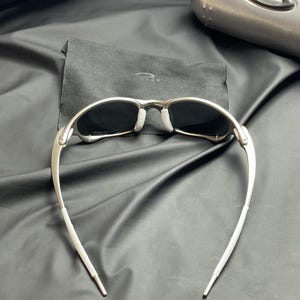May include: Silver-framed sunglasses with dark lenses and white nose pads. The sunglasses have a sleek, modern design with curved temples. A black cloth case is behind the sunglasses. The background is a black, textured surface.
