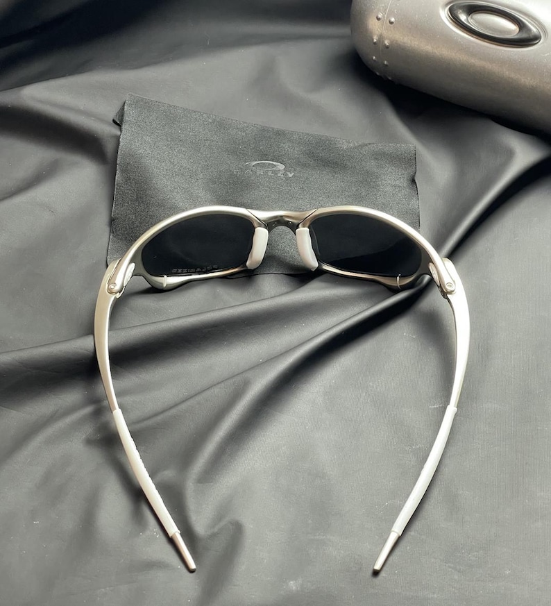 May include: Silver-framed sunglasses with dark lenses and white nose pads. The sunglasses have a sleek, modern design with curved temples. A black cloth case is behind the sunglasses. The background is a black, textured surface.