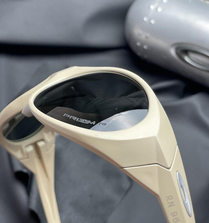 Puede incluir: Gafas de sol beige con lentes oscuras. Las gafas de sol tienen un dise&ntilde;o deportivo con el texto "PRIZM" visible en la lente. La montura es de color beige claro y las patillas tienen el texto "RN 96".