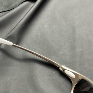 May include: Close-up of a pair of silver-toned sunglasses with black lenses. The frame has a sleek, modern design with a white accent on the temple. The text "04-114A 56-21 135" is printed on the temple.