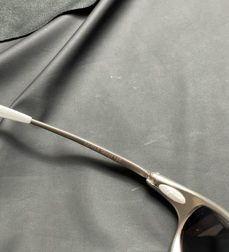 May include: Close-up of a pair of silver-toned sunglasses with black lenses. The frame has a sleek, modern design with a white accent on the temple. The text "04-114A 56-21 135" is printed on the temple.