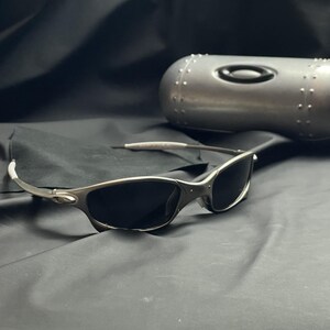 May include: Silver-framed sunglasses with dark lenses rest on a black surface, next to a matching silver case. The sunglasses have a sleek, modern design. The case is oval-shaped with a black logo.