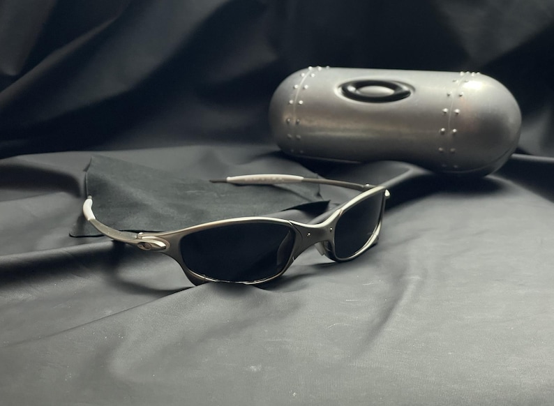 May include: Silver-framed sunglasses with dark lenses rest on a black surface, next to a matching silver case. The sunglasses have a sleek, modern design. The case is oval-shaped with a black logo.